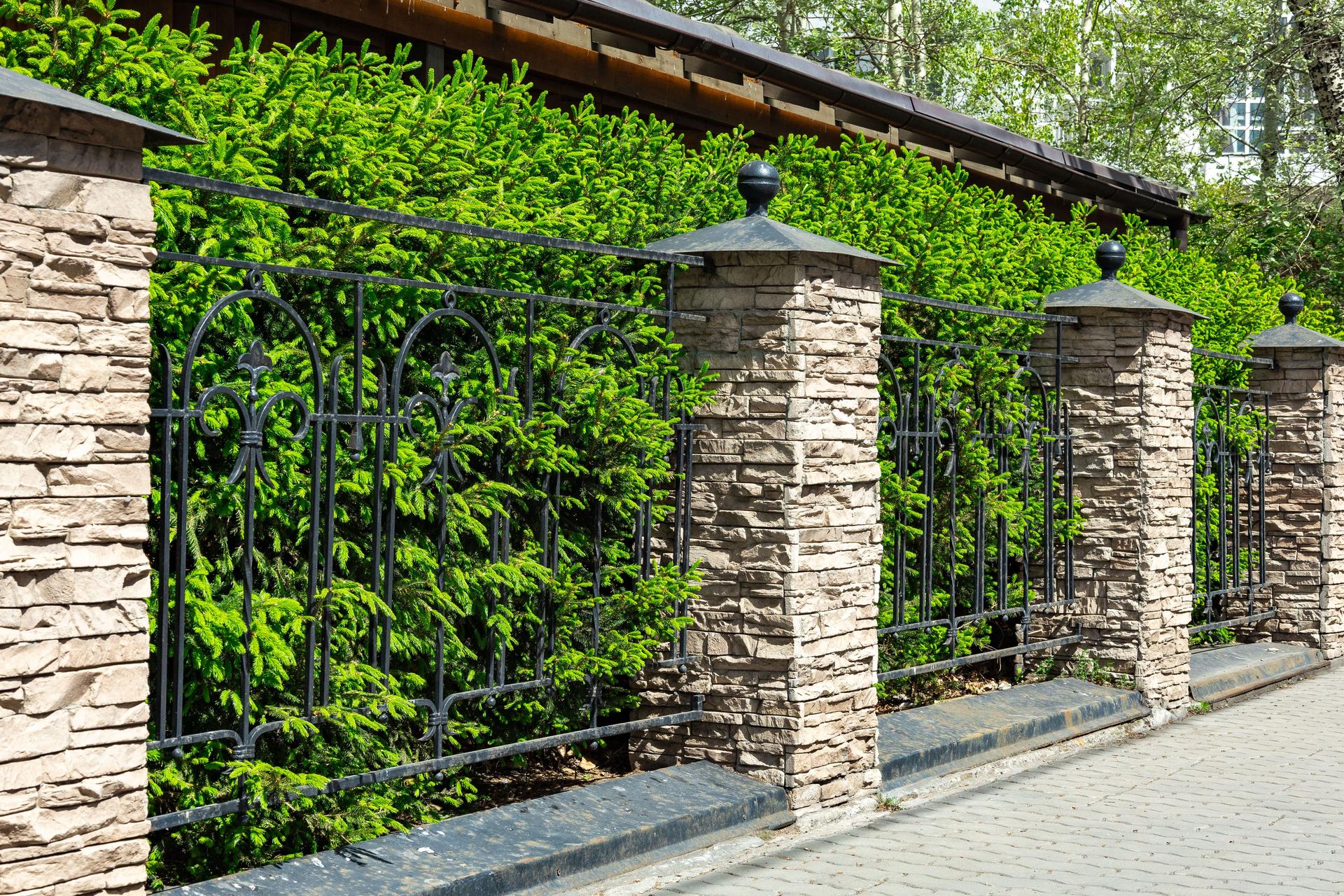 Elegant stone and wrought iron fence bordering a lush green hedge on a sunny day in a peaceful residential area